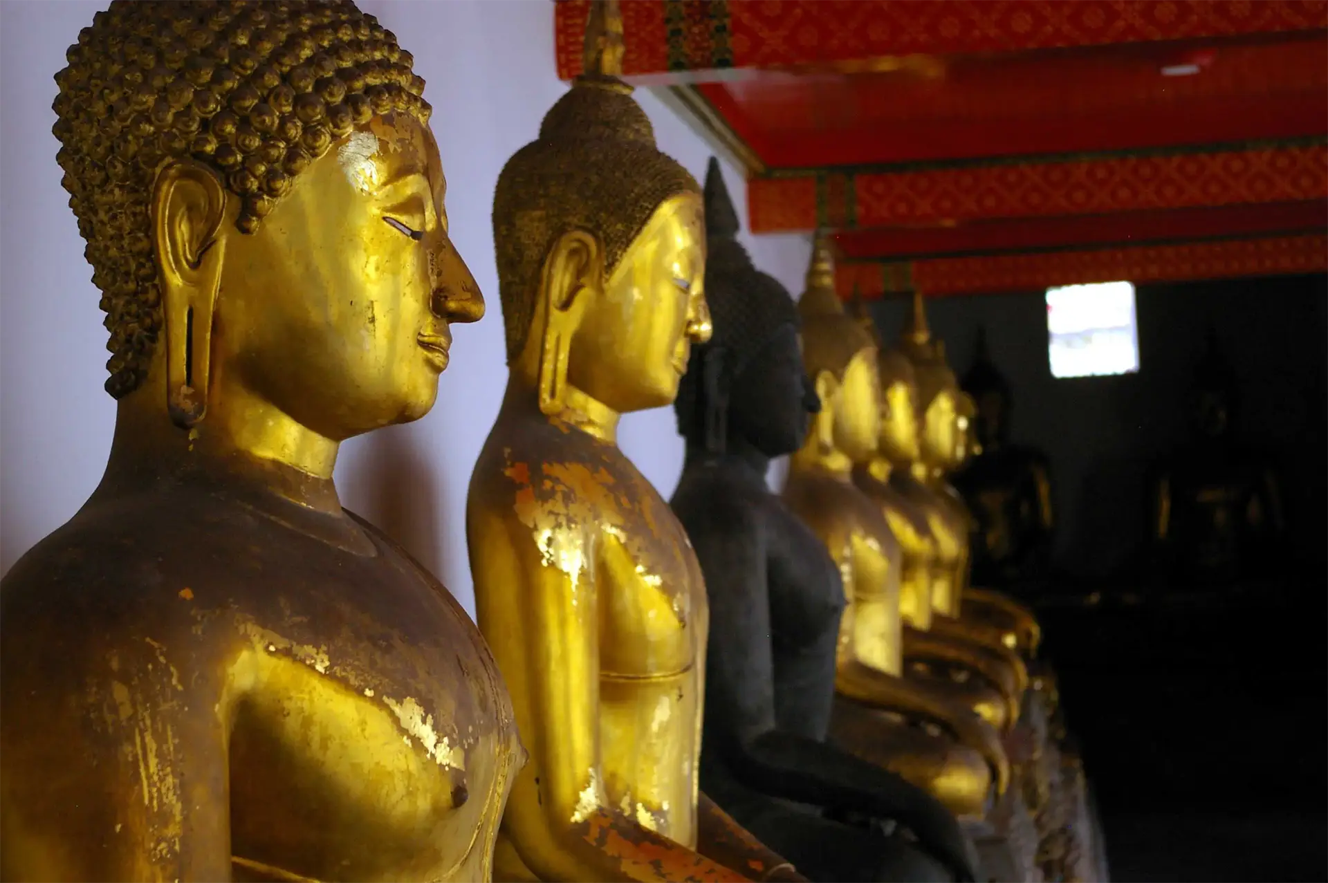 Rangée de bouddhas dorés et patinés alignés dans la galerie du Wat Pho à Bangkok, sous un plafond rouge