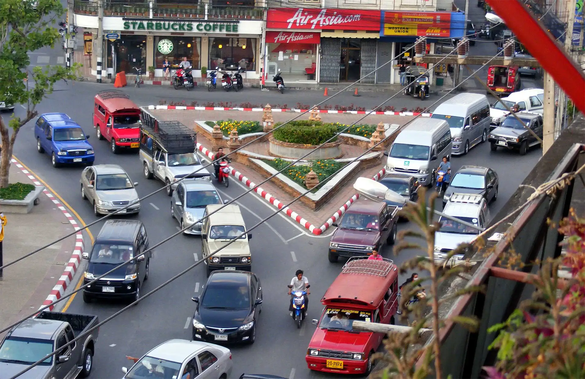 Carrefour animé de Chiang Mai vu d’un balcon, songthaews rouges, voitures et scooters autour d’un rond-point