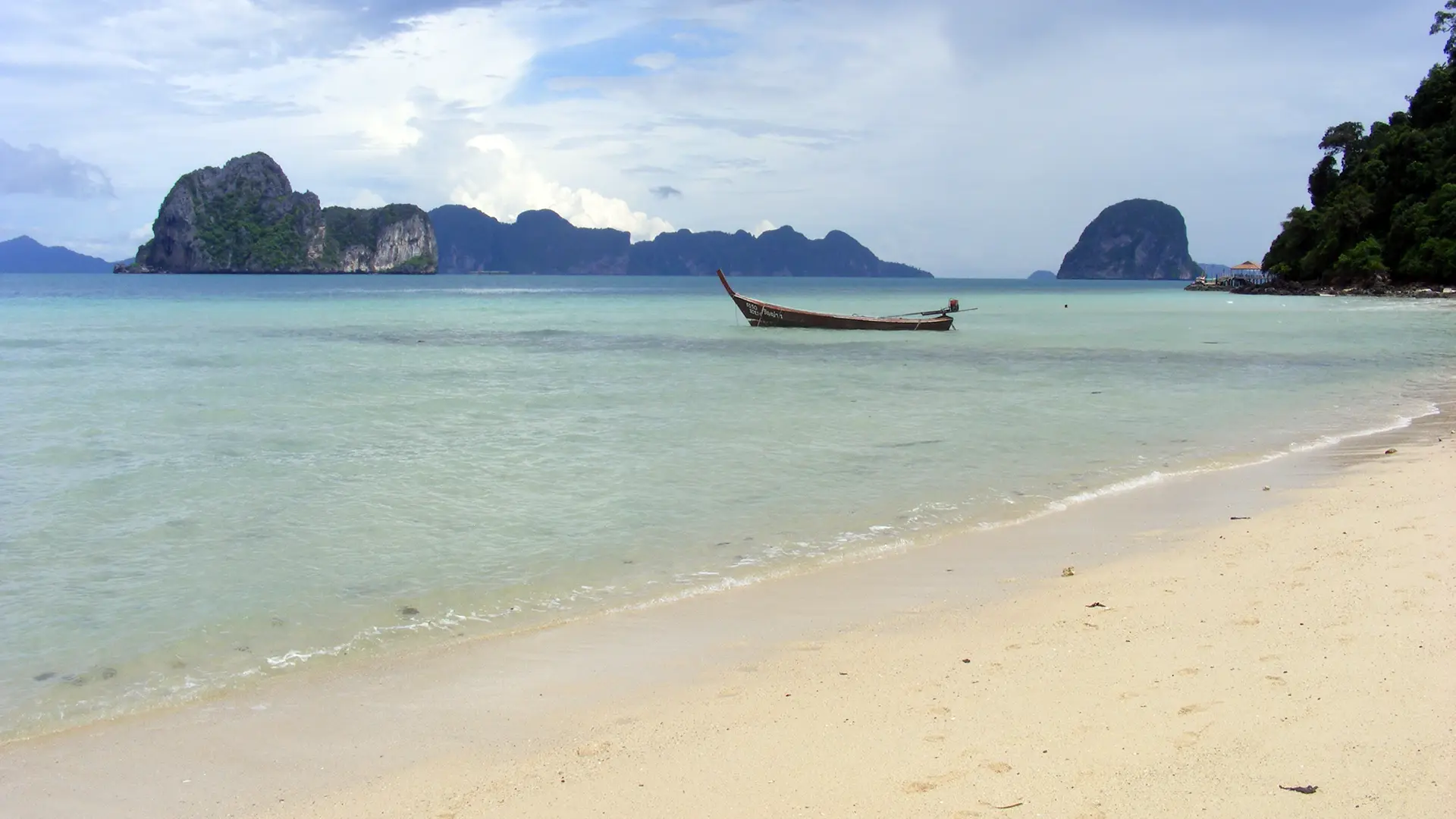 Plage de sable blanc de Koh Ngai, eau turquoise et bateau à longue queue face aux pitons calcaires de la mer d’Andaman