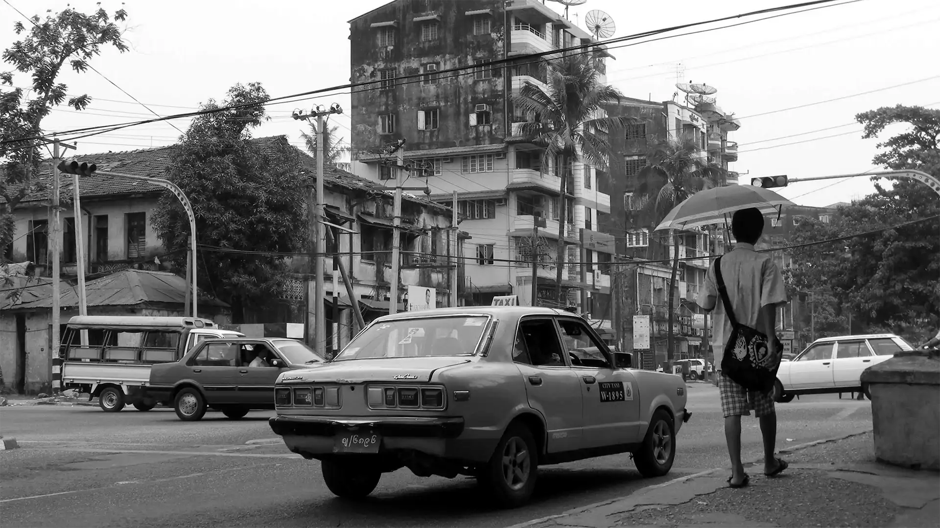 Rue de Rangoun en noir et blanc, un vieux taxi et un passant en longyi abrité sous un parapluie